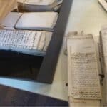 Ein Tisch, auf dem viele alte Manuskripte liegen. Rechts oben ist ein Teil einer Macbook-Tastatur zu sehen.
